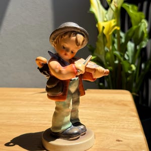Könnte beinhalten: Eine Keramikfigur einer jungen Person mit grauem Hut, roter Jacke und hellgrüner Hose, die eine kleine Geige spielt. Die Figur trägt eine dunkle Tasche über der Schulter und steht auf einem runden Sockel, beleuchtet von hellem Sonnenlicht.