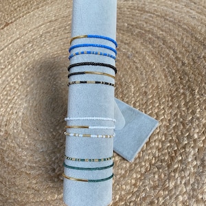 Peut inclure: Un présentoir de bracelets en perles avec des accents dorés. Les bracelets sont empilés sur un support gris et présentent différentes couleurs, notamment le bleu, le noir, le blanc et le vert.