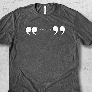 May include: Dark grey heather t-shirt with the word "quote" in white, between two white quotation marks. The shirt is made of a soft material and has a crew neck. The shirt is a casual style.
