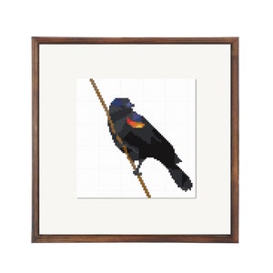 May include: A framed cross-stitch artwork depicting a black bird with a red and orange patch on its wing, perched on a brown branch.