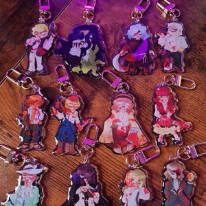 Peut inclure: Une collection de 13 porte-clés en acrylique colorés représentant différents personnages avec des éclaboussures de sang. Les personnages sont représentés dans un style cartoon et affichent diverses expressions. Les porte-clés sont attachés à des anneaux métalliques dorés.