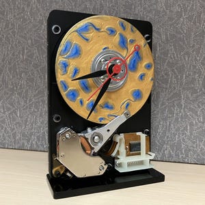 May include: A unique clock crafted from a repurposed hard drive. The clock face is a metallic gold color with blue accents. Black clock hands and a red second hand are visible. The clock is mounted on a black base.