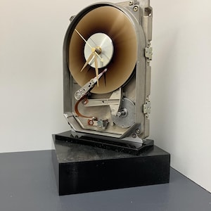 May include: A wall clock repurposed from a computer hard drive, featuring a circular brown platter as the clock face, gold-colored hands, and internal mechanical components mounted on a rectangular black base.