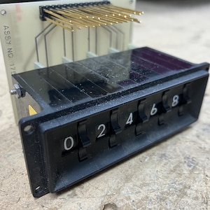 May include: A black electromechanical thumbwheel counter module featuring a series of rotating numbered wheels displaying digits 0, 2, 4, 6, and 8. The unit is attached to a beige printed circuit board with a row of gold-colored metal connector pins.