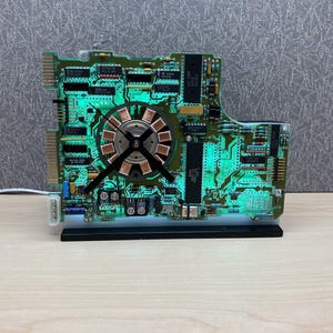 May include: A unique clock crafted from a green circuit board, featuring a copper-colored center and black clock hands. The clock is illuminated with a green glow and sits on a black base, showcasing a blend of technology and design.