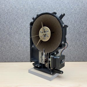 May include: A unique, steampunk-style clock with a circular, tan-colored face and gold-colored hands. The clock's mechanism is visible, featuring a black frame, silver components, and a small motor. It sits on a rectangular, gray base.