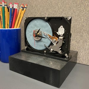 May include: A unique clock made from a repurposed hard drive, featuring a light blue disc and clock hands. It sits on a black rectangular base, next to a blue mug filled with pencils.