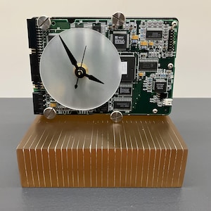 May include: A desk clock constructed from a green circuit board featuring a circular silver face with black hands. The clock is mounted on a rectangular, amber-colored, translucent base with vertical ribbed detailing and four silver-toned screws.