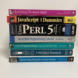 May include: A stack of nine books about programming languages, including JavaScript, Perl, and UNIX. The books are all different colors and have different titles. The books are stacked on top of each other on a white surface.