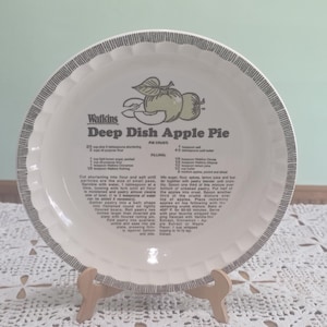 May include: A white ceramic pie dish with a fluted edge and a dark gray striped rim. The center features a green apple illustration, the text Watkins Deep Dish Apple Pie, and a printed recipe with instructions for crust and filling preparation.