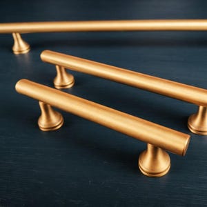 May include: Three cylindrical cabinet pulls with a brushed gold finish. Each handle features a long, straight bar supported by two flared, circular bases. The hardware is arranged in a staggered formation, showcasing the smooth, metallic texture.