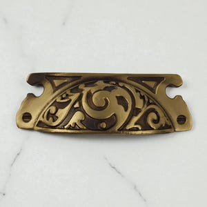 May include: A brass drawer pull with a semi-circular center featuring an embossed scrollwork pattern. The hardware has a horizontal rectangular shape with notched sides and two circular screw holes on the far left and right ends for mounting.