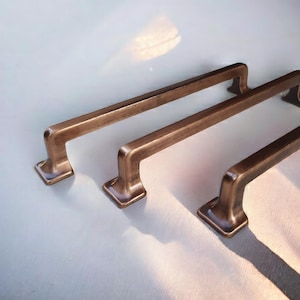 Puede incluir: Tres tiradores de cajón de metal con acabado en bronce cepillado. Cada manija presenta un diseño de barra larga y recta con dos bases de montaje cuadradas en los extremos. Los herrajes tienen un tono metálico cálido con reflejos oscuros.
