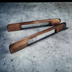 May include: Two elongated cabinet pulls with a brushed copper finish. Each handle features a flat, rectangular bar design with rounded ends and two mounting points, showcasing a metallic texture and a warm, reddish-brown color throughout.
