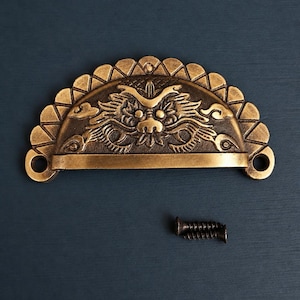 May include: A semi-circular brass-toned metal drawer pull featuring an embossed dragon face design with a scalloped top edge and two screw holes on the sides. A single small metal screw is positioned below the handle for installation purposes.