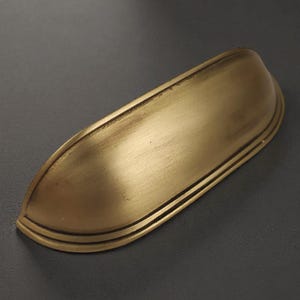 Könnte beinhalten: Ein halbkreisförmiger Muschelgriff für Schubladen mit gebürstetem Messing-Finish. Der Beschlag verfügt über eine glatte, abgerundete Oberfläche und einen gestuften Rand an der Basis, in einem metallischen Goldton mit dezenter Patina.