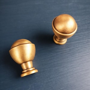 May include: Two brass cabinet knobs with a brushed metallic finish. Each knob features a rounded, dome-shaped top that transitions into a tapered, cylindrical base with a circular mounting plate at the bottom, showcasing a warm, golden hue.