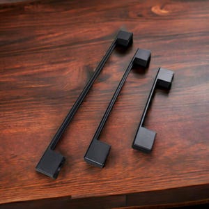 May include: Three matte black cabinet pulls of varying lengths arranged diagonally. Each handle features a long, slender bar with a square-shaped mounting post at each end, providing a clean, geometric look for furniture or cabinetry hardware.