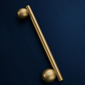 May include: A gold-toned metal cabinet handle featuring a long, cylindrical bar attached to two spherical ends. The hardware has a smooth, matte finish and is positioned diagonally across the frame, showcasing a clean, geometric design.