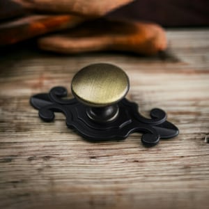 May include: A cabinet knob featuring a round, brushed brass-toned top attached to a matte black metal backplate. The backplate has an elongated, symmetrical shape with curved, scroll-like decorative edges extending from the central post.