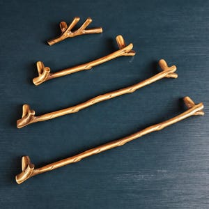 May include: Four gold-toned metal cabinet pulls shaped like tree branches. Each handle features a textured, organic form with small twig-like protrusions at the ends and along the length, varying in size from a small knob to long pulls.