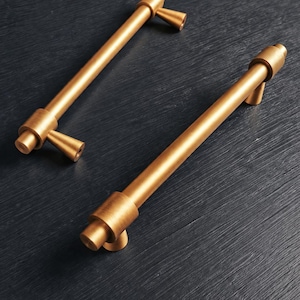 May include: Two brushed brass cabinet pulls with a cylindrical bar design and conical mounting posts. Each handle features a smooth, matte metallic finish with a warm golden tone, showcasing a clean, linear structure for furniture or cabinetry hardware.