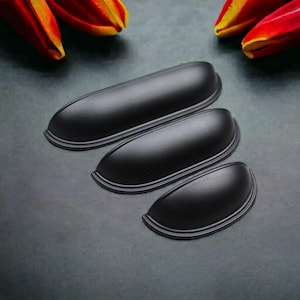 Könnte beinhalten: Drei mattschwarze Muschelgriffe aus Metall in unterschiedlichen Längen, die diagonal angeordnet sind. Jeder Griff hat eine glatte, abgerundete, halbzylindrische Form mit einer geschwungenen Basis und einem definierten Rand für Möbel.