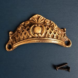 Può includere: Una maniglia per cassetto in metallo color ottone a forma di semicerchio, caratterizzata da un motivo a griglia decorato e un elemento circolare centrale. L'accessorio presenta due fori di montaggio laterali e due piccole viti nere.
