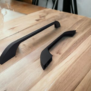 Könnte beinhalten: Zwei mattschwarze Schrankgriffe mit geschwungenem, länglichem Design. Der größere Griff befindet sich links, während der kleinere Griff rechts liegt und zwei kreisförmige Schraublöcher an der Basis seines Befestigungspunktes für die Montage zeigt.