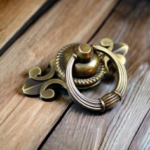 May include: A brass-toned metal drawer pull featuring an ornate, elongated backplate with scrollwork details and a circular, hinged ring handle. The hardware has a weathered, antique finish with visible dark highlights in the recessed areas.