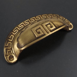 May include: A semi-circular brass drawer pull featuring an antique finish and a raised Greek key pattern along the curved edge. The center of the handle displays a square spiral motif, with two screw holes located at the ends of the hardware piece.