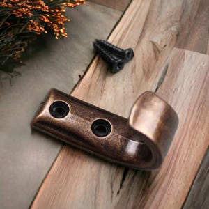 Può includere: Un gancio da parete singolo con una finitura in bronzo scuro anticato e due fori circolari per le viti di montaggio. Il gancio presenta un design curvo a forma di J ed è accompagnato da una vite in metallo nero con gambo filettato.