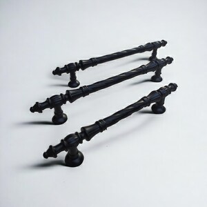 May include: Three matte black cabinet pulls arranged in a diagonal row. Each handle features a central twisted metal bar design with decorative finials at both ends and two circular mounting bases that connect the handle to the cabinet surface.