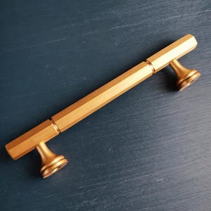 Op de afbeelding: Een goudkleurige metalen meubelgreep met een lange, zeshoekige stang en twee uitlopende, ronde montagevoeten. Het beslag heeft een gladde, matte afwerking en een recht, langwerpig ontwerp dat aan beide uiteinden op de voeten is bevestigd.