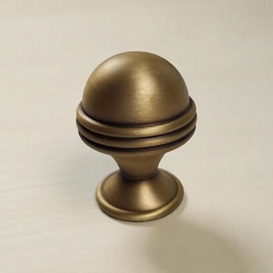 May include: A brass-toned metal cabinet knob featuring a smooth, rounded dome top, a central section with two horizontal recessed grooves, and a flared circular base. The finish has a brushed, matte appearance with subtle dark highlights.