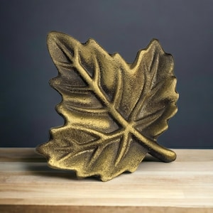 May include: A decorative leaf-shaped dish with a textured, metallic gold finish. The object features a prominent central vein and branching veins extending toward the jagged, irregular edges of the leaf, creating a realistic botanical appearance.