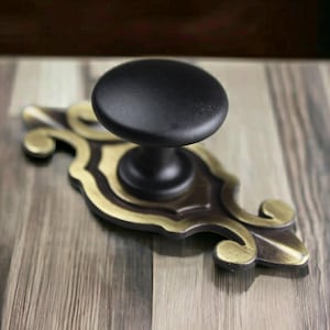 May include: A matte black round cabinet knob attached to an elongated, decorative backplate. The backplate features a metallic antique brass finish with ornate, curved edges and a pointed tip, providing a contrast to the simple black knob.