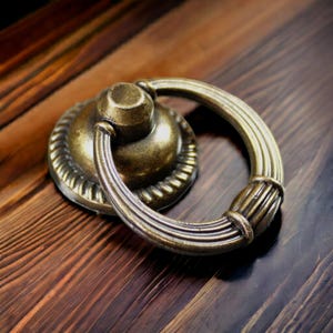 Puede incluir: Un tirador de cajón circular de metal en tono latón con una base de montaje central, un borde con textura de cuerda y un asa de anillo acanalada y articulada. El herraje tiene un acabado antiguo desgastado con tonos oscuros.