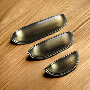 Könnte beinhalten: Drei Muschelgriffe für Schubladen in einem dunklen, gebürsteten Antikmessing-Finish. Jeder Griff weist ein glattes, geschwungenes, halbkreisförmiges Design mit einer vertieften Mitte und einem erhabenen Rand auf, angeordnet nach absteigender Größe.