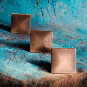 May include: Three square cabinet knobs with a brushed copper finish. Each knob features a flat, slightly raised center surface with smooth, beveled edges, arranged in a diagonal line to showcase the metallic texture and warm brown tones.