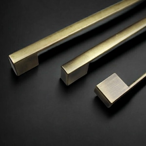 Peut inclure: Trois poignées de placard linéaires avec une finition en laiton brossé et un profil rectangulaire. Chaque poignée présente une construction en métal solide avec une texture mate lisse et des bords nets et précis, disposées en diagonale.