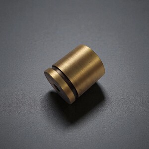 May include: A cylindrical brass knob with a brushed metallic finish. The hardware features a flat circular face with a central hole and a distinct recessed groove near the base, showcasing a smooth, solid construction and a warm golden tone.
