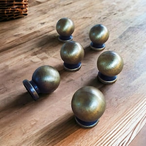 May include: Six spherical cabinet knobs featuring a metallic gold finish on the rounded tops and a dark purple base. Each knob has a smooth, polished surface that reflects light, creating a consistent appearance across the set of hardware pieces.