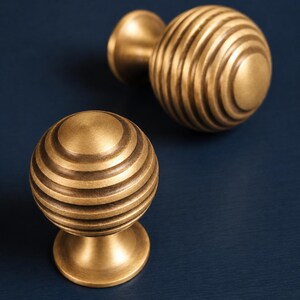 May include: Two spherical cabinet knobs made of brushed brass with a series of horizontal circular grooves encircling the main body. Each knob features a smooth, flat circular top and a flared base that tapers inward toward the center point.