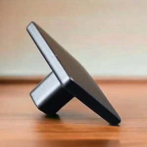 May include: A matte black cabinet knob featuring a square-shaped front face and a cylindrical base. The hardware is positioned at an angle, showcasing its solid construction and smooth, dark finish, designed for attachment to furniture or cabinetry doors.