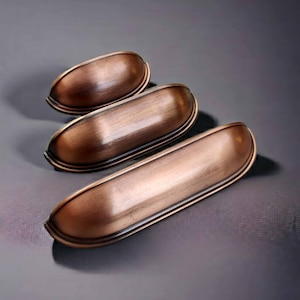 Könnte beinhalten: Drei Muschelgriffe in gebürsteter Kupferoptik, die diagonal von klein nach groß angeordnet sind. Jeder Griff hat eine glatte, abgerundete, halbzylindrische Form mit einem ausgeprägten Rand an der Basis und einer metallischen Textur.