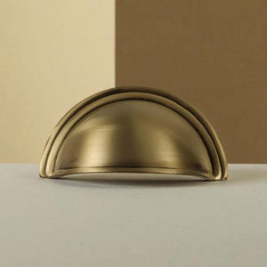 Op de afbeelding: Een halfronde komgreep voor laden met een geborstelde messing afwerking. Het beslag heeft een gladde, afgeronde koepelvorm met een platte onderkant en een verhoogd randdetail langs de gebogen bovenkant van het metalen object.