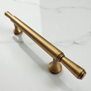 Könnte beinhalten: Ein gebürsteter goldener Möbelgriff mit einer langen zylindrischen Stange und zwei ausgestellten Befestigungspfosten. Der Griff weist dezente ringförmige Details an den Enden und eine glatte, matte Metalloberfläche auf.