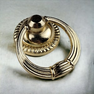 Peut inclure: Une poignée de tirage annulaire en métal doré dotée d'une base circulaire avec une bordure texturée en forme de corde. L'anneau mobile présente un design cannelé et nervuré avec une bande centrale lisse, offrant une prise fonctionnelle.