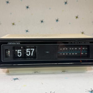 May include: Vintage Soundesign AM/FM digital clock radio. The clock displays "5:57" in large white numbers. The radio has a cream-colored top with a black front panel, and a tuning dial. The radio has a retro aesthetic.